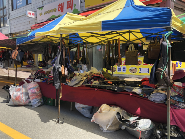 Ein Markt mit Zelten, die Taschen, Hüte und andere Gegenstände präsentieren, vor Gebäuden mit Fenstern und Texttafeln im Hintergrund.