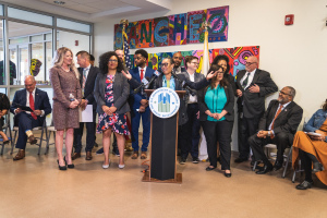 Gruppe von Menschen vor einem Podium mit einem Mikrofon und einer Tafel, auf der "Sancho City Councilman Joins City Council" steht, mit sitzenden und stehenden Teilnehmern, Flaggen, einem Feuerlöscher und Deckenleuchten im Hintergrund.