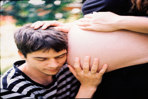Ein Mann und eine Frau stehen nebeneinander, der Mann berührt zärtlich den Bauch der Frau, mit einem unscharfen Hintergrund.