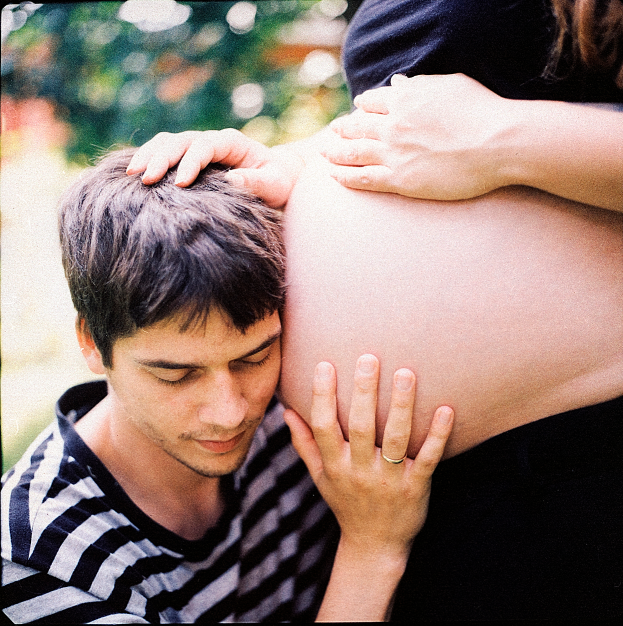 Ein Mann und eine Frau stehen nebeneinander, der Mann berührt zärtlich den Bauch der Frau, mit einem unscharfen Hintergrund.