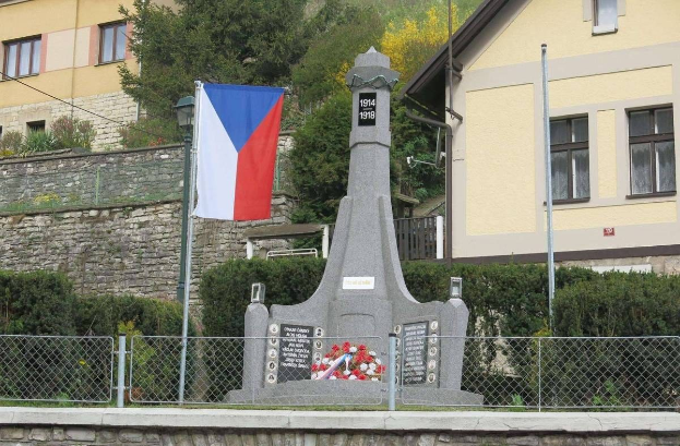 Ein Denkmal mit einer Flagge darauf, umgeben von einem Zaun, Pflanzen, Bäumen und einer Wand, vor einem Gebäude, gewidmet denen, die ihr Leben während des Zweiten Weltkriegs verloren haben, mit eingravierter Schrift.
