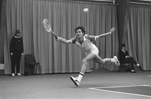 Ein Mann schwingt einen Tennisschläger gegen einen Ball, mit zwei stehenden und zwei sitzenden Personen im Hintergrund und einem Vorhang als Rahmen.