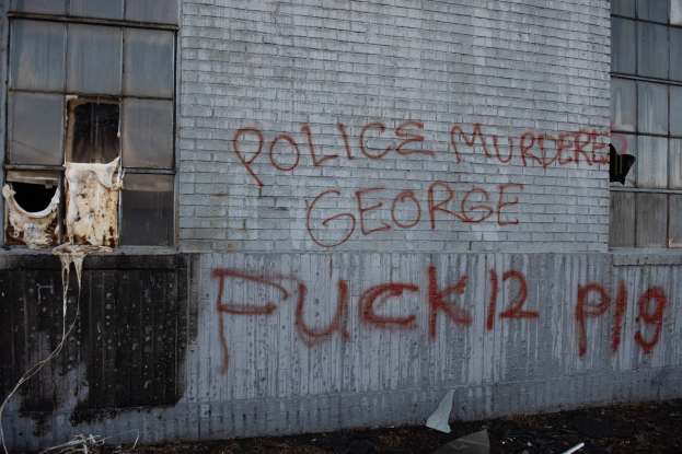 Graffiti an einer Gebäudewand mit der Aufschrift "Polizei ermordet George Fuck 12 P19" und Fenstern und verstreuten Gegenständen davor.