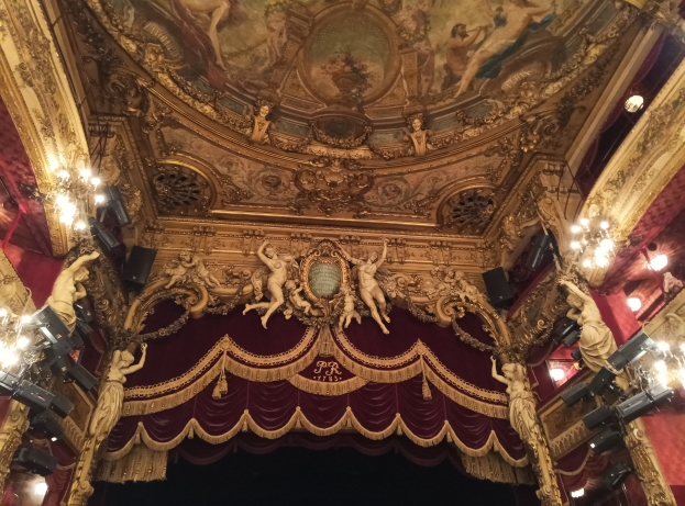 Theaterinnenraum mit roter Gardine, Kronleuchtern, Deckenplastiken, Lichtern und einem Gemälde, das eine Bühne und einige Personen unten zeigt.