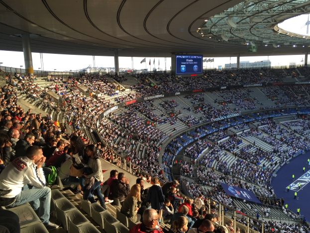 Eine große Menschenmenge sitzt im Allianz Stadion in München, Deutschland, und schaut ein Füpballspiel, mit einer Bühne auf der rechten Seite, Flaggen, Masten und einem Bildschirm im Hintergrund und dem Himmel oben sichtbar.