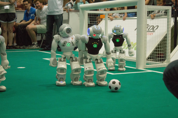 Eine Gruppe von Robotern, die auf einem grünen Feld Fußball spielen, mit einem Ball in der Mitte, einem Netz auf der rechten Seite und Zuschauern mit Kameras im Hintergrund.