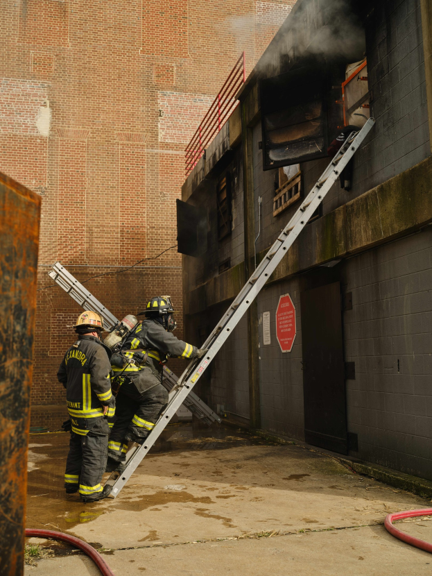 Feuerwehrleute mit Helmen, die an einem raucherfüllten Gebäude eine Leiter hochsteigen, mit einem Metallobjekt links und Rohren auf dem Boden.