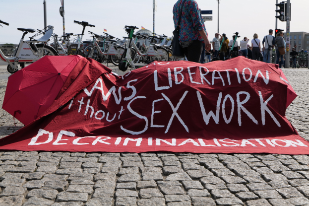Eine Gruppe von Menschen demonstriert auf einer Stadtstraße, hält Schirme und zeigt ein rotes Tuch mit der Aufschrift "Befreiung ohne Entkriminalisierung der Sexarbeit" auf dem Boden, mit Fahrrädern, Verkehrsinfrastruktur und Gebäuden im Hintergrund.
