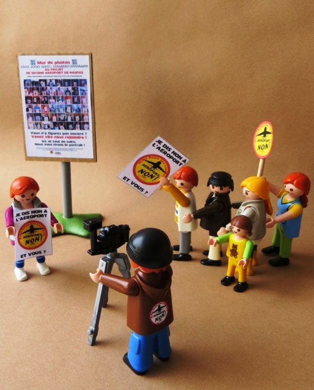 Menschliche Puppen mit Plakaten, eine Kamera auf einem Stativ und eine Hintergrundtafel mit Bildern und Text.