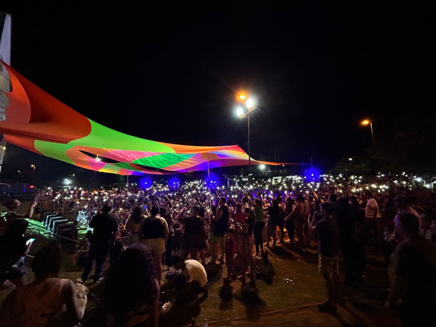 Eine große Menschenmenge steht nachts vor einem Zelt, das von bunten Lichtern beleuchtet wird und von beleuchteten Pfählen umgeben ist, mit einem Gebäude auf der linken Seite und dem Nachthimmel im Hintergrund während des 2018er Kunstfestivals.