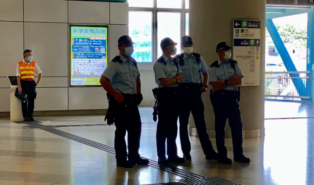 Eine Gruppe von maskierten Polizisten vor einem Gebäude mit Glastüren, Informationsschildern, einer Säule und einem Geländer, mit Bäumen und Fahrzeugen im Hintergrund