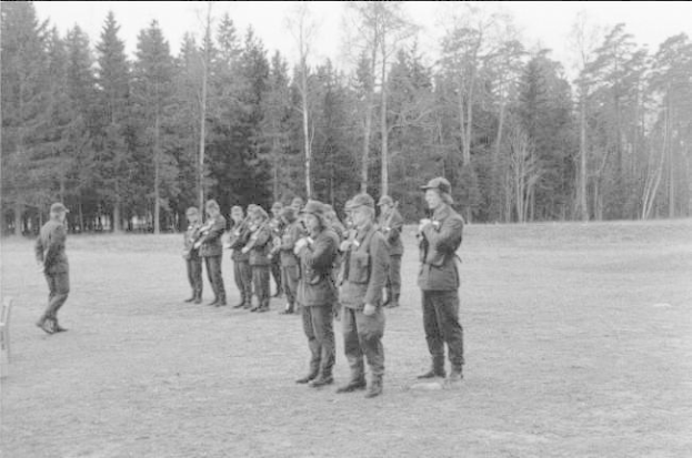 Schwarz-weißes Bild einer Gruppe von Männern in Militäruniformen mit Mützen und Gewehren, die in einem Feld mit Bäumen und einem klaren Himmel stehen, wahrscheinlich während eines Trainingsübungs.