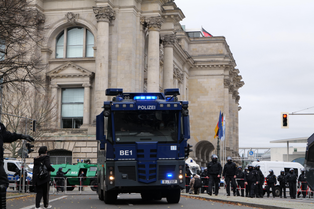 Eine Gruppe von Polizeibeamten steht vor einem großen Gebäude mit Fenstern, Säulen und Bögen, mit Fahrzeugen auf der Straße, einer Person mit einer Kamera auf der linken Seite und einem klaren blauen Himmel mit Bäumen, Ampeln, Pfählen und Fahnen im Hintergrund.