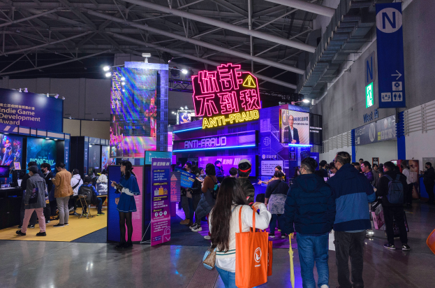 Eine Gruppe von Menschen steht vor einem Messestand, einige halten Taschen, mit Schildern, die 'Indie Game Development Award 2019' anzeigen, Bildschirmen, Lichtern und anderen Gegenständen im Hintergrund und einer Wand auf der rechten Seite.
