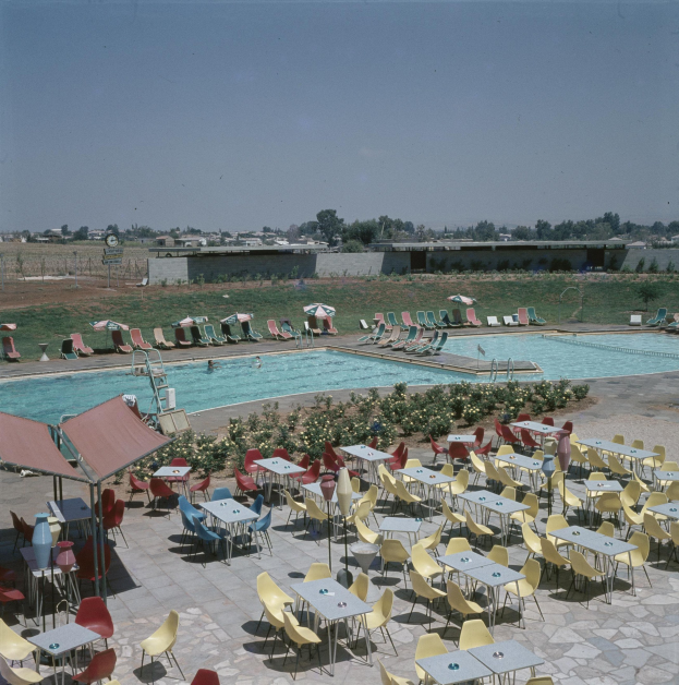 Ein Freibad mit Tischen, Stühlen und Sonnenschirmen, umgeben von Blumenpflanzen, einem Zaun, einer Wand, Bäumen, Gebäuden und einer bewölkten Himmel.