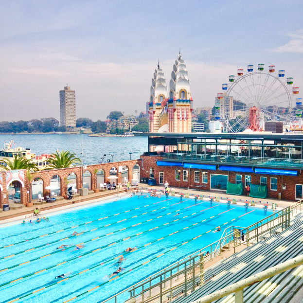 Ein großes Schwimmbad mit schwimmenden Menschen, umgeben von Geländern und Bänken, mit einem riesigen Riesenrad, Gebäuden, Bäumen und einem bewölkten Himmel im Hintergrund.