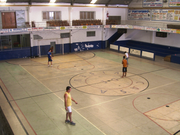 Eine Gruppe von Menschen, die Badminton in einer Turnhalle mit einem Netz in der Mitte spielt, eine Wand mit Fenstern und Türen im Hintergrund, Banner mit Text auf der rechten Seite und Lampen an der Decke.