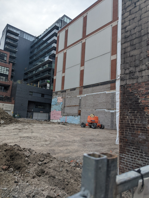 Baustelle mit einem großen Gebäude im Hintergrund, das abgerissen wird, einem Fahrzeug auf dem Boden, einem Metallzaun und einem bewölkten Himmel.