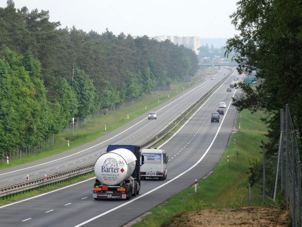 Ein gro├če Lastwagen fahrt auf einer von beiden Seiten mit B├Ąumen ges├Ąumten Autobahn, mit einem Zaun und Gras auf der rechten Seite und Geb├Ąuden im Hintergrund unter einem klaren blauen Himmel.