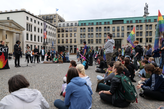 Eine Gruppe von Menschen, die auf dem Boden vor einer Menge sitzen, die Fahnen und Plakate hält, mit einer Person, die in ein Mikrofon spricht, einer Statue und Gebäuden im Hintergrund während einer Anti-Schwulen-Demo in Berlin.