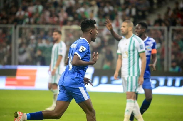 Fußballspieler in einem blauen und weißen Trikot, der nach einem Tor vor jubelnden Fans feiert, umgeben von Teamkollegen in weißen und grünen Trikots und einer jubelnden Menge im Hintergrund.