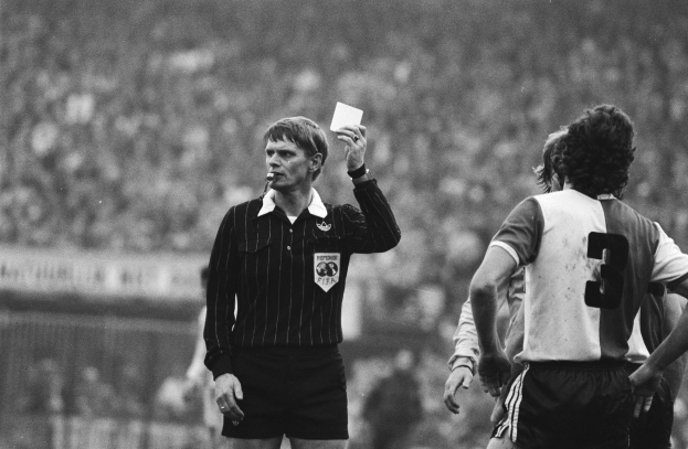 Schwarzes und weißes Bild eines Schiedsrichters, der eine Rote Karte vor einer Menge von Fußballspielern hält, mit einem Zaun im Hintergrund.