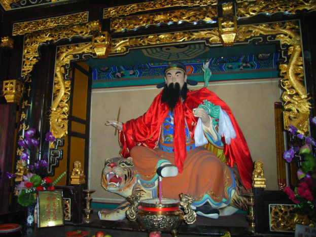 Eine Statue eines chinesischen Mönchs mit buntem Stoff auf einem Tiger sitzt in einem Bogen mit goldenen Verzierungen, vorne ein Blumenstrauß und in der Mitte eine Schüssel.