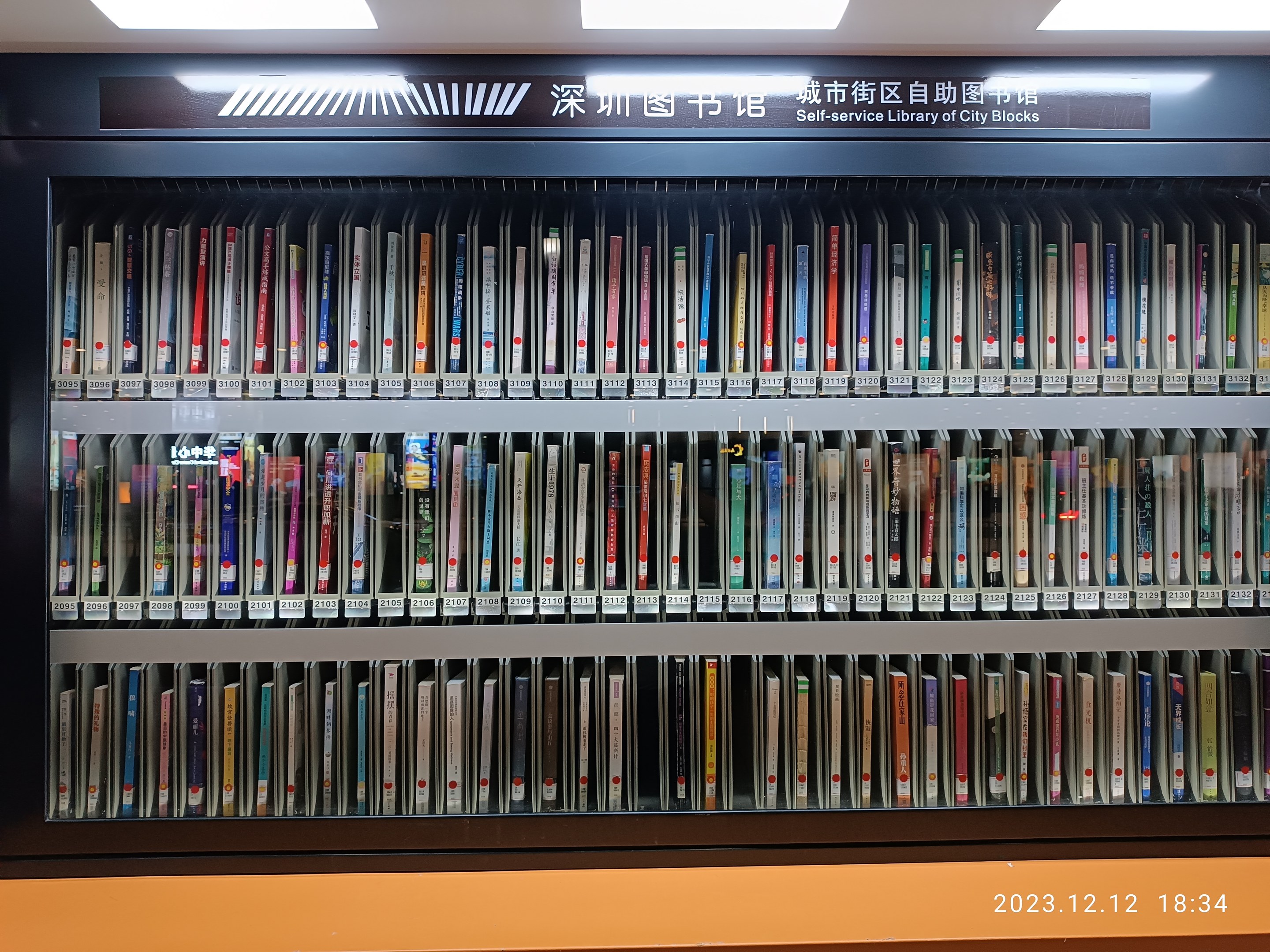 Eine gut beleuchtete Bibliothek mit hohen Bücherregalen voller Bücher, ein sichtbares Textbrett und ein Wasserzeichen unten, das an eine Selbstbedienungsbibliothek in Stadtblocks erinnert.