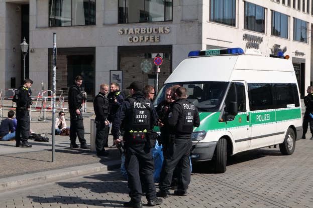 Eine Gruppe von Polizeibeamten steht vor einem Starbucks-Café mit einem Van auf der rechten Seite und einigen Menschen auf der linken Seite, mit einem Gebäude, einem Schild, einem Laternenmast und einem Zaun im Hintergrund.