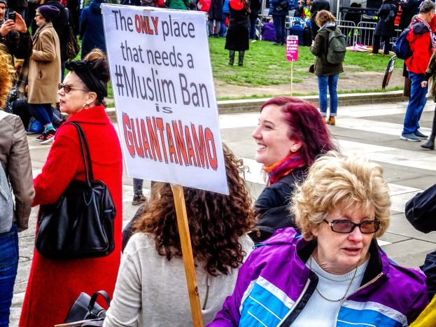 Eine Gruppe von Menschen steht auf dem Boden, einige halten Taschen, eine Frau hält ein Schild mit der Aufschrift "Der einzige Ort, an dem ein Muslimverbot gebraucht wird, ist Guantanamo". Im Hintergrund sehen wir einen Metallzaun, Gras, ein Schild mit Text, einige Pfosten, ein Gebäude mit Fenstern und Bäume.