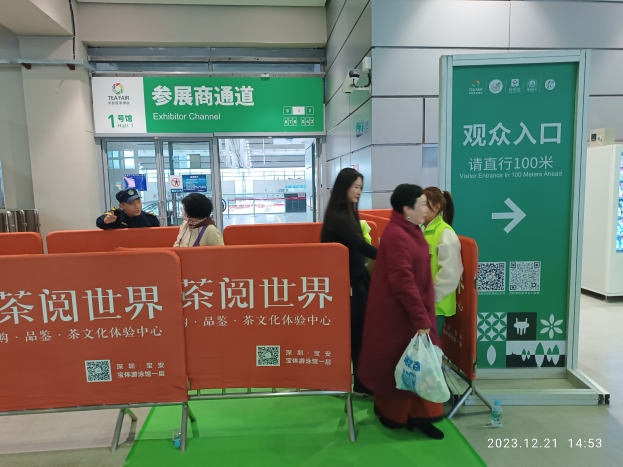 Eine Gruppe von Menschen steht vor einem grünen Teppich auf einer Tee-Messe in China, mit Schildern, einer Flasche auf dem Boden, einem Display, Metallstangen, Schautafeln, einer Überwachungskamera und einer Decke mit Deckenleuchten.