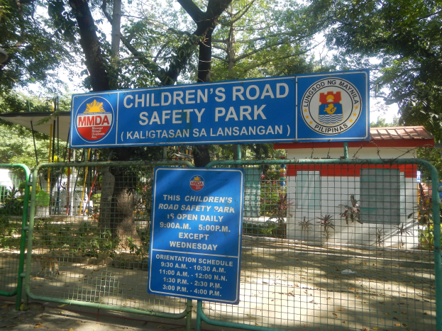 Metallzaun um einen Kinder-Verkehrssicherheitspark mit zwei blauen Informationsschildern, einem kleinen Haus, Pflanzen, Bäumen und einem bewölkten Himmel.