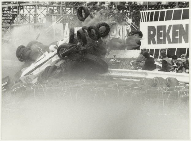 Schwarz-weiß-Foto eines verunglückten Rennwagens, umgeben von Reifen, mit einer Gruppe von Menschen rechts und einer Tafel mit Text im Hintergrund.