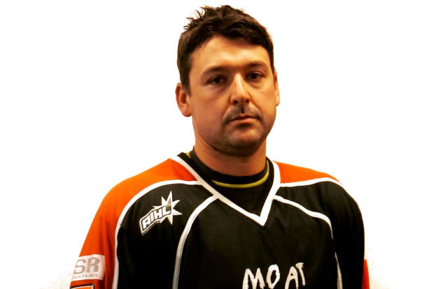 Mann in schwarzem und orangefarbenem Hockey-Jersey mit ernstem Gesichtsausdruck, Hände verschränkt, kurzes dunkles Haar und Kinnbart vor weißem Hintergrund.