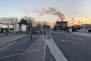 Strassenszene mit Verkehrskegeln, einem Bus, Laternenmasten, Bäumen, Gebäuden, Kränen und Rauch, der aus einem entfernten Gebäude aufsteigt.