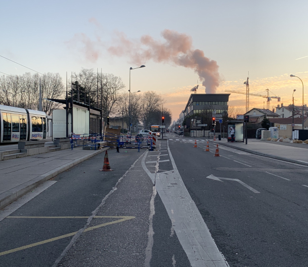 Strassenszene mit Verkehrskegeln, einem Bus, Laternenmasten, Bäumen, Gebäuden, Kränen und Rauch, der aus einem entfernten Gebäude aufsteigt.