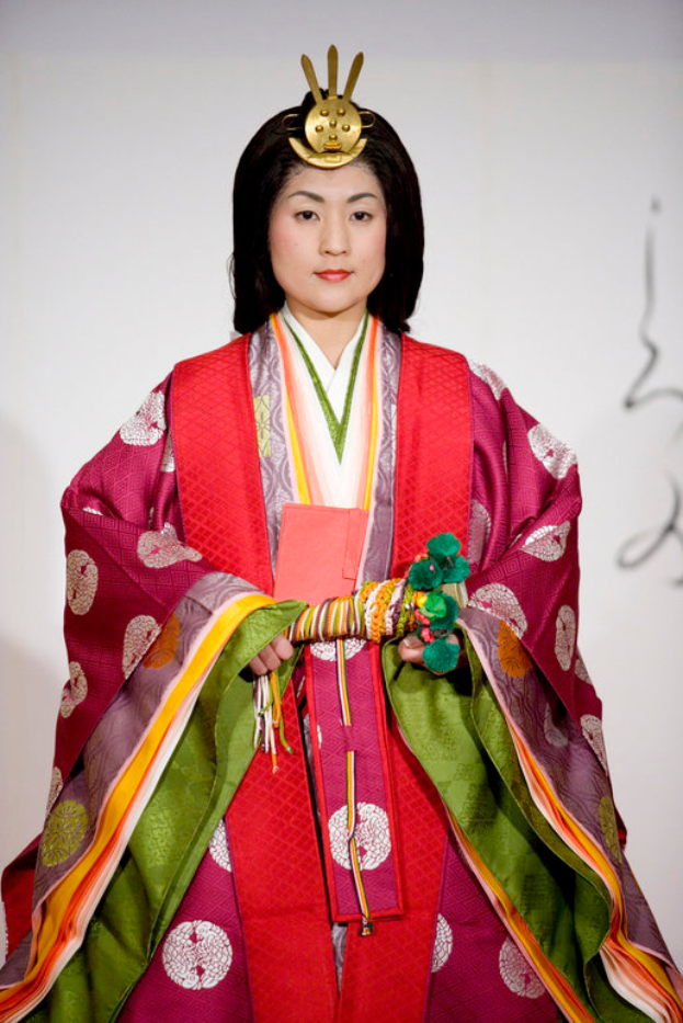 Eine Frau in einem traditionellen japanischen Kimono, das ein leuchtendes rotes und grünes Kleid mit kunstvollen Mustern zeigt, steht selbstbewusst auf einem Laufsteg, ihr Haar ist ordentlich zu einem Knoten gebunden.