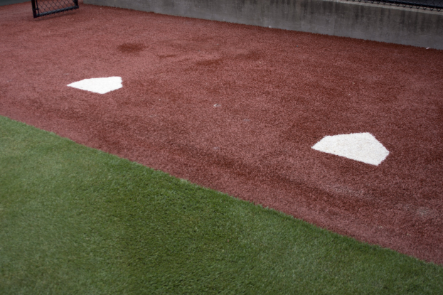 Baseballfeld mit Kunststoffrasen, einer Umzäunung, Home Plate in der Mitte und einer Wand im Hintergrund.