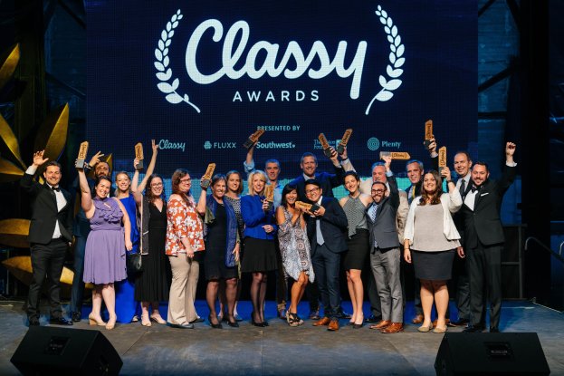 Gruppe von Menschen auf einer Bühne, die Auszeichnungen halten, mit Lautsprechern unten und einem Bildschirm im Hintergrund, der "Classy Awards 2019" anzeigt.