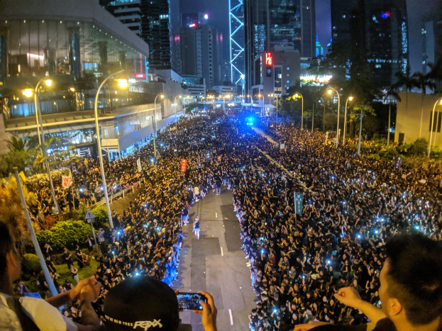 Eine Menschenmenge steht nachts auf einer Straße, beleuchtet von Straßenlaternen, einige halten Handys hoch während einer Protestaktion in Hong Kong.