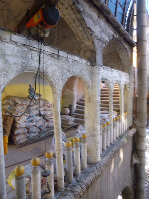 Altes Gebäude mit Bogengang und Säulen wird renoviert, mit verstreuten Werkzeugen, Materialien und Gegenständen, darunter Taschen und eine Maschine mit Drähten auf dem Boden.