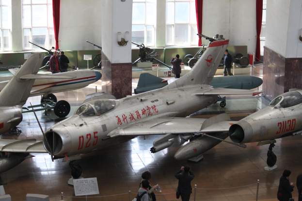 Eine Gruppe von Kampfflugzeugen in einem Museum ausgestellt, mit Besuchern, die hinter Barrieren beobachten, informativen Schildern und architektonischen Elementen.