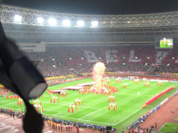 Ein Stadion mit Flutlicht, Eisen Gittern, Zuschauern, stehenden Personen, Werbetafeln, Bildschirmen, Sicherheitskameras, Flaggen und Dekorationen.