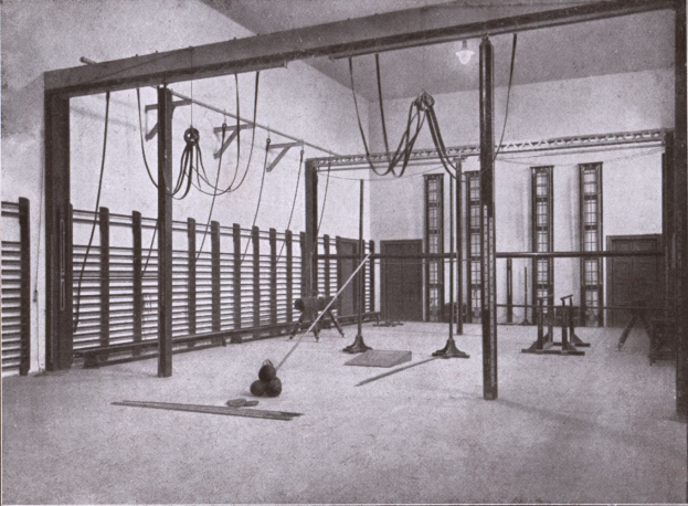 Ein Schwarz-Weiß-Foto eines Fitnessstudios mit verschiedenen Geräten wie Stangen und Seilen auf dem Boden, Fenstern und einer Wand im Hintergrund und Text unten.
