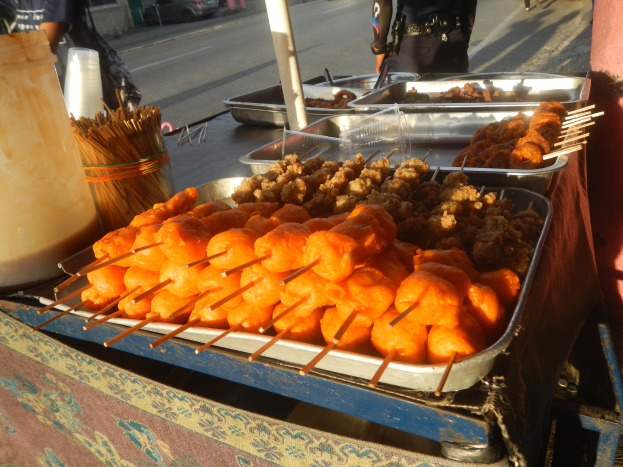 Ein Straßenhändler verkauft Essen von einem Tisch mit Tabletts voller Gegenstände, Zahnstochern, einem Eimer und anderen Gegenständen, mit Menschen, Fahrzeugen, Pfählen, Gebäuden, Bäumen und einem klaren blauen Himmel im Hintergrund.