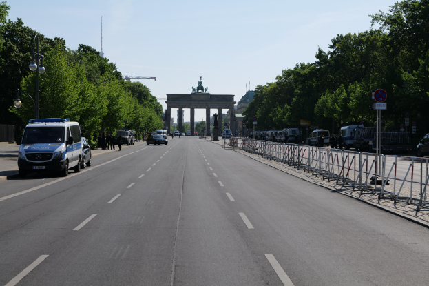 Ein Polizeiwagen steht auf der Seite einer vielbefahrenen Straße vor dem Brandenburger Tor in Berlin, Deutschland, mit Barrieren, Schildern, Bäumen und Laternen im Hintergrund und einer bewölkten Himmel.