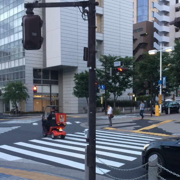 Scooter auf einer Straße mit Füßgängern im Hintergrund, eine Ampel im Vordergrund und Gebäude mit Bäumen dahinter.