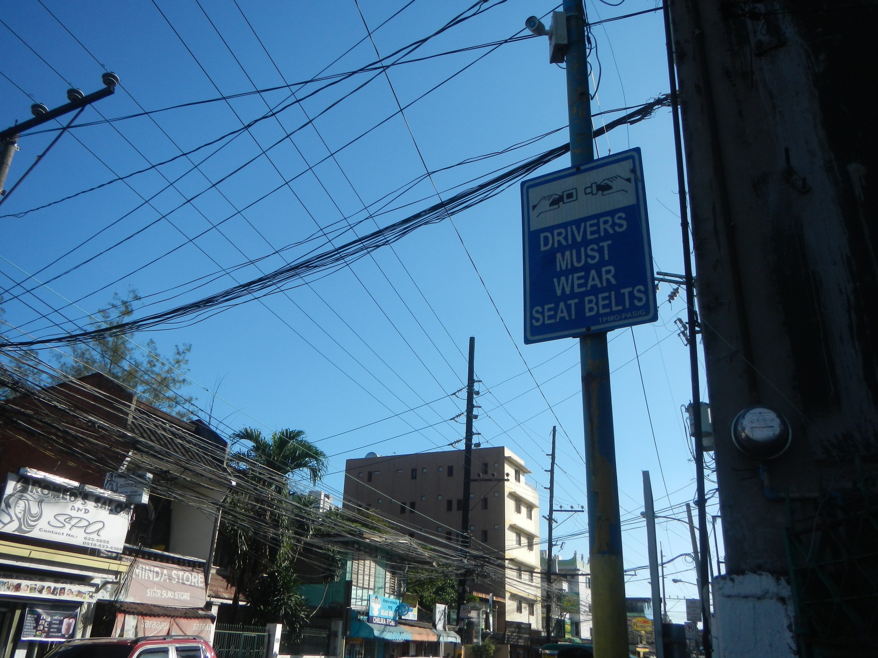 Stadtstraße mit fahrenden Autos, Strommasten mit Drähten, Gebäuden mit Fenstern, Bäumen, Namensschildern und einem "Fahrer müssen sich anschnallen"-Schild an einem Strommast vor einem sichtbaren Himmel.