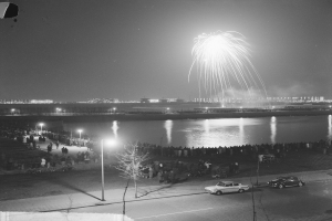 Ein Schwarz-Weiß-Foto einer Menge, die Feuerwerk über Wasser beobachtet, mit Fahrzeugen, Laternenpfählen, Bäumen und Gebäuden im Hintergrund.