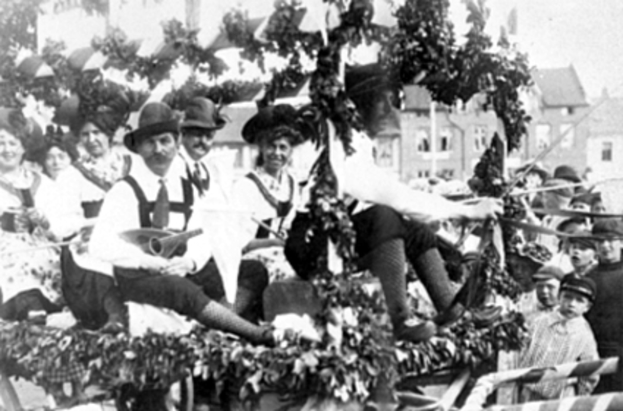 Eine Gruppe von Menschen in traditioneller bayrischer Tracht fährt auf einem mit Blättern geschmückten Festwagen mit, einige spielen Musikinstrumente, im Hintergrund sind Gebäude und ein klarer Himmel zu sehen, dargestellt in Schwarz-Weiß.